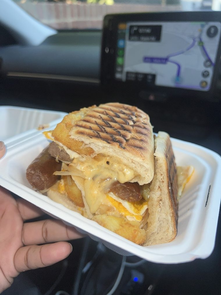 A hand holding an open takeaway box with a toasted panini inside a car